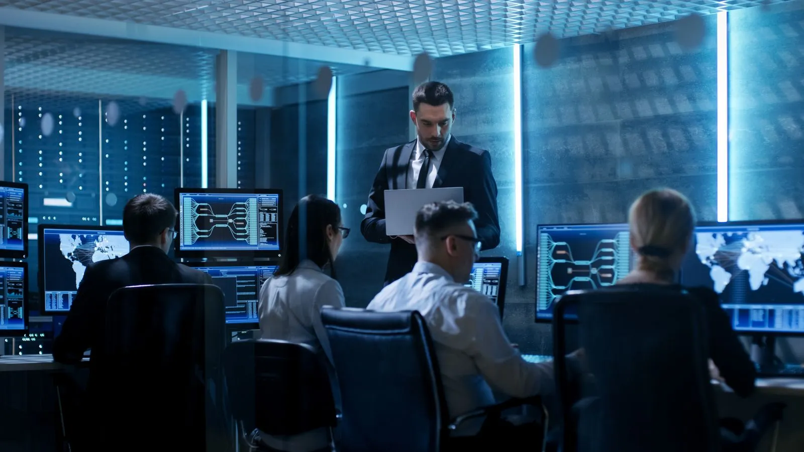 A team of professionals working intently at computer stations in a modern, high-tech control room, with one person standing and reviewing data on a laptop.